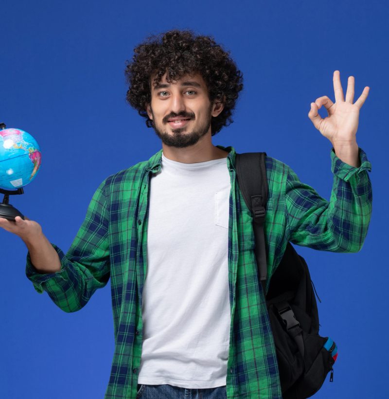 front-view-male-student-green-checkered-shirt-wearing-black-backpack-holding-little-globe-smiling-blue-wall