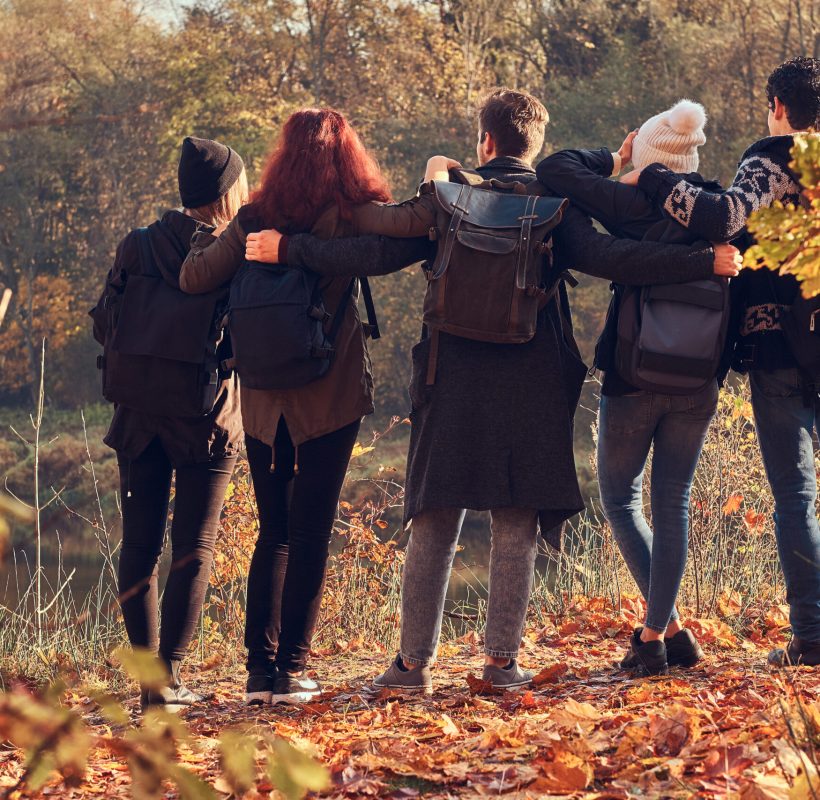 Travel, hiking, adventure concept. Back view of young friends hugging together and looking at the lake in beautiful autumn forest.